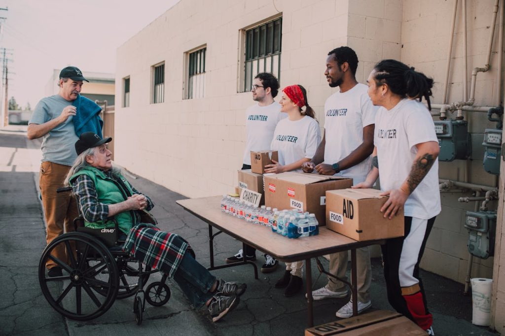 pexels photo 6646914 Volunteers assist a senior adult in a wheelchair by distributing aid packages outdoors.